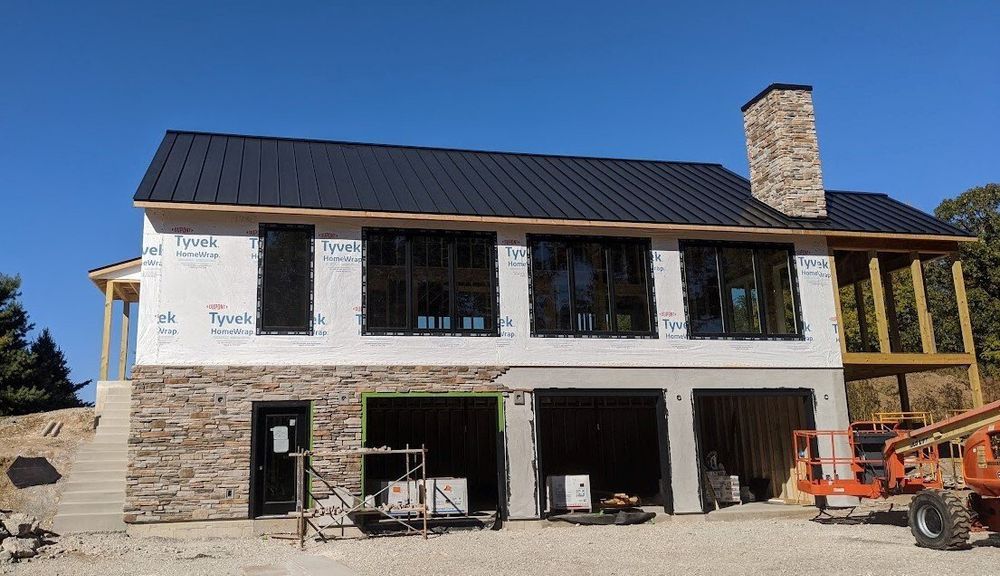 Two-story house under construction with stone facade, black windows, and a black roof against a blue sky. Two-story house under construction with stone base, black roof, and construction equipment in front