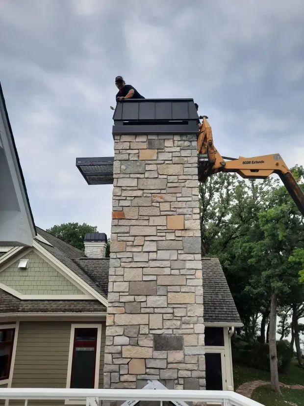 Two workers on a stone chimney roof, one using a lift against a cloudy sky