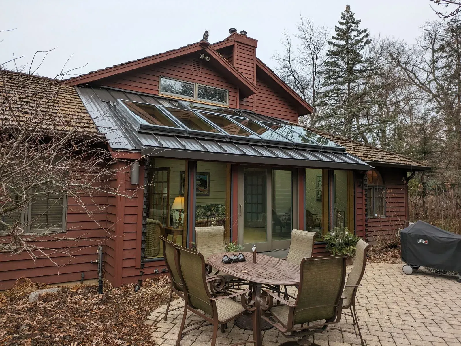 Red house with a metal roof extension, patio furniture, and glass doors. Overcast day. Red wooden house patio with glass awning, outdoor table, chairs, and grill on a stone terrace
