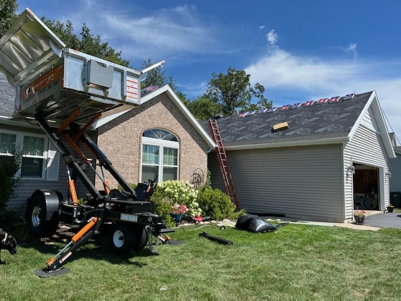 Truck-mounted lift raising debris from a house roof beside a garage in a suburban yard.