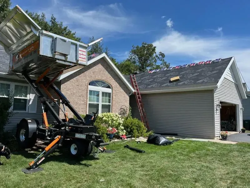 A house with a mechanical lift in the yard, likely for roofing work. Ladder, and debris are visible. Blue sky. Moving truck lifting a furniture-filled box into a suburban garage driveway under a blue sky