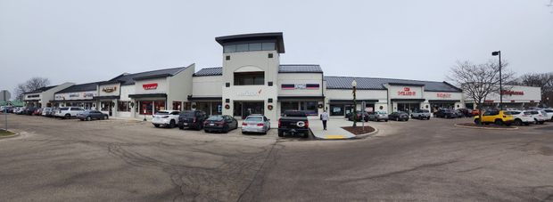 Wide parking lot outside a strip mall with storefronts, parked cars, and a cloudy sky