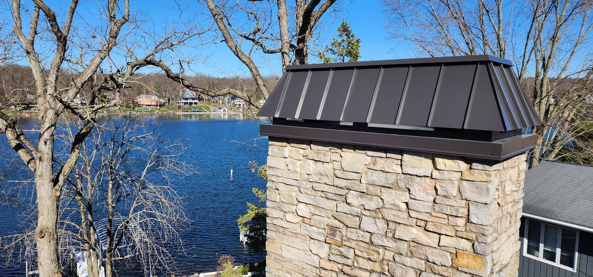 Stone chimney with a dark metal cap, blue water and trees in the background.