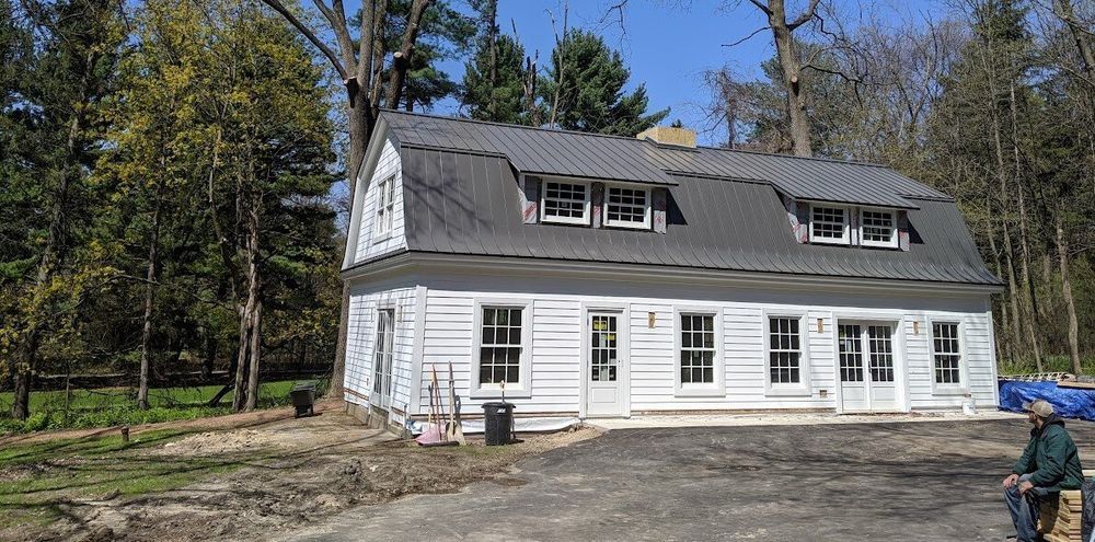 White two-story house with gray roof and dormer windows in a wooded yard, person at right edge