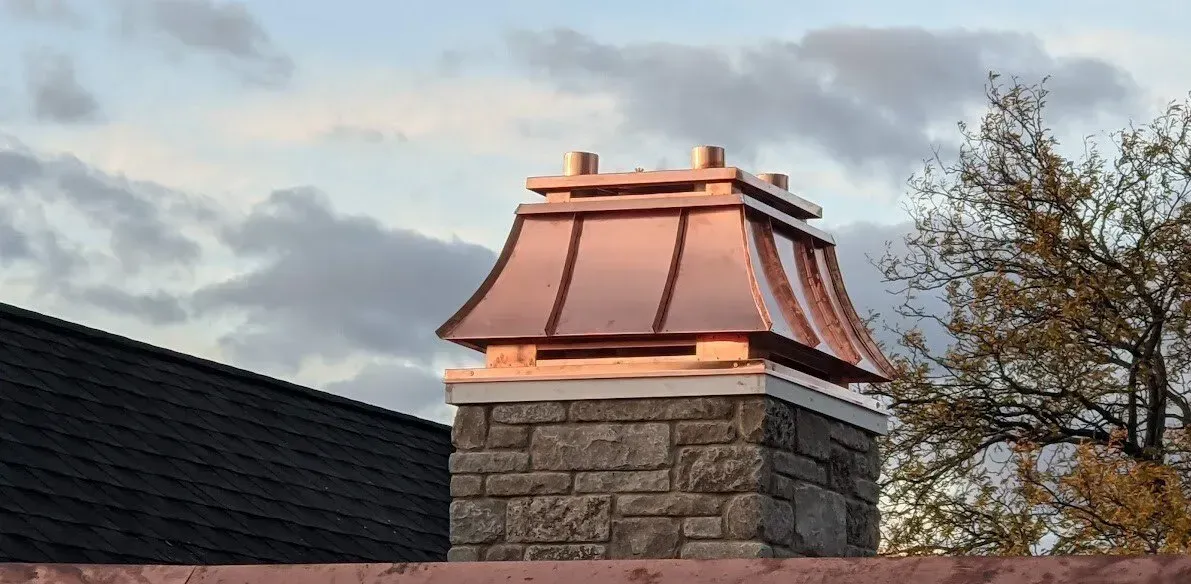 Stone chimney with copper roof cap against a cloudy sky. Copper-roofed stone chimney against a cloudy sky beside a dark roof and tree branches