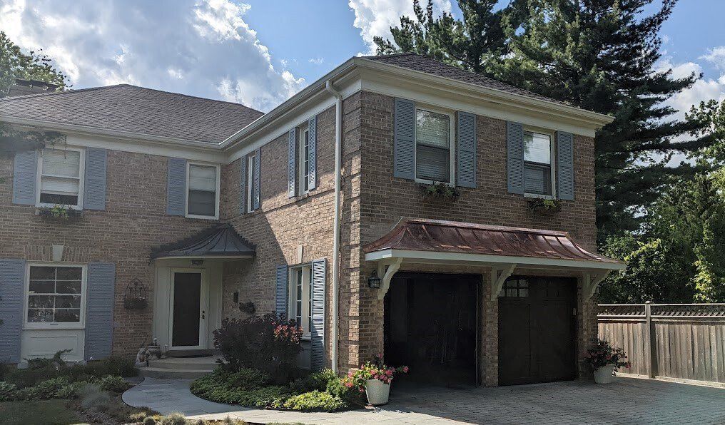 Two-story brick house with attached garage, dark shingled roof, and front walkway under a partly cloudy sky