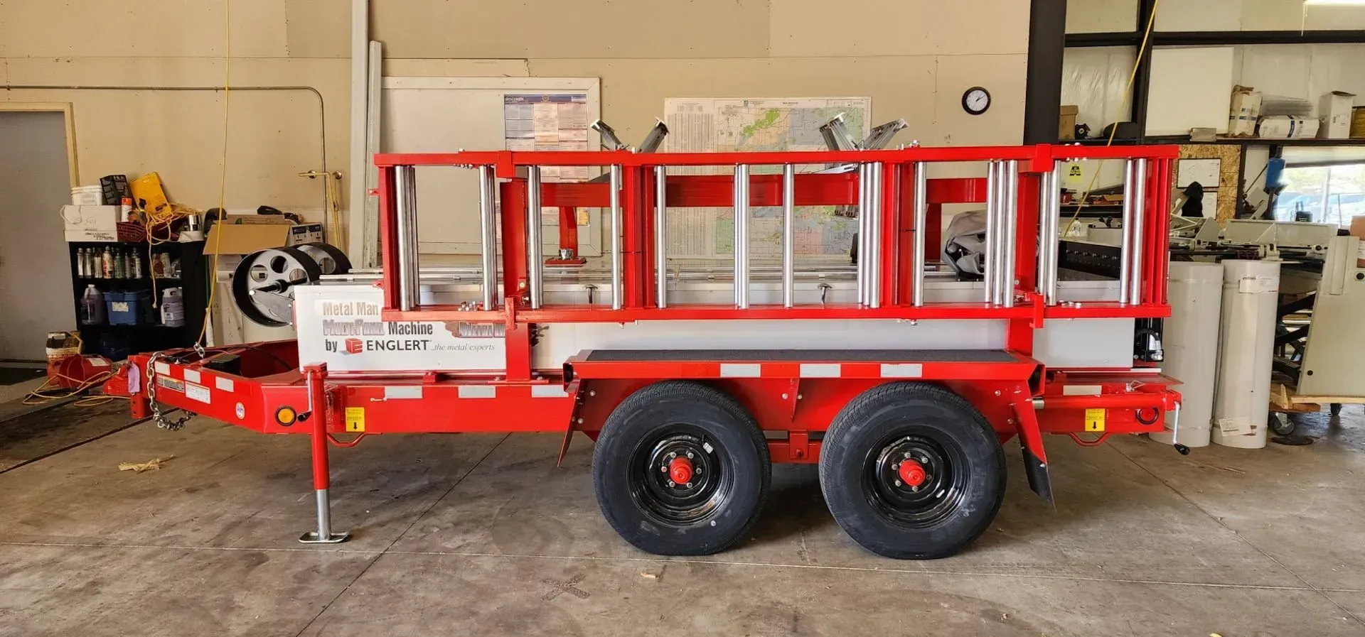 Red and white lift machine on a trailer. Two large black tires, ladder-like lift structure. Red utility trailer with mesh sides and black wheels parked inside a workshop.