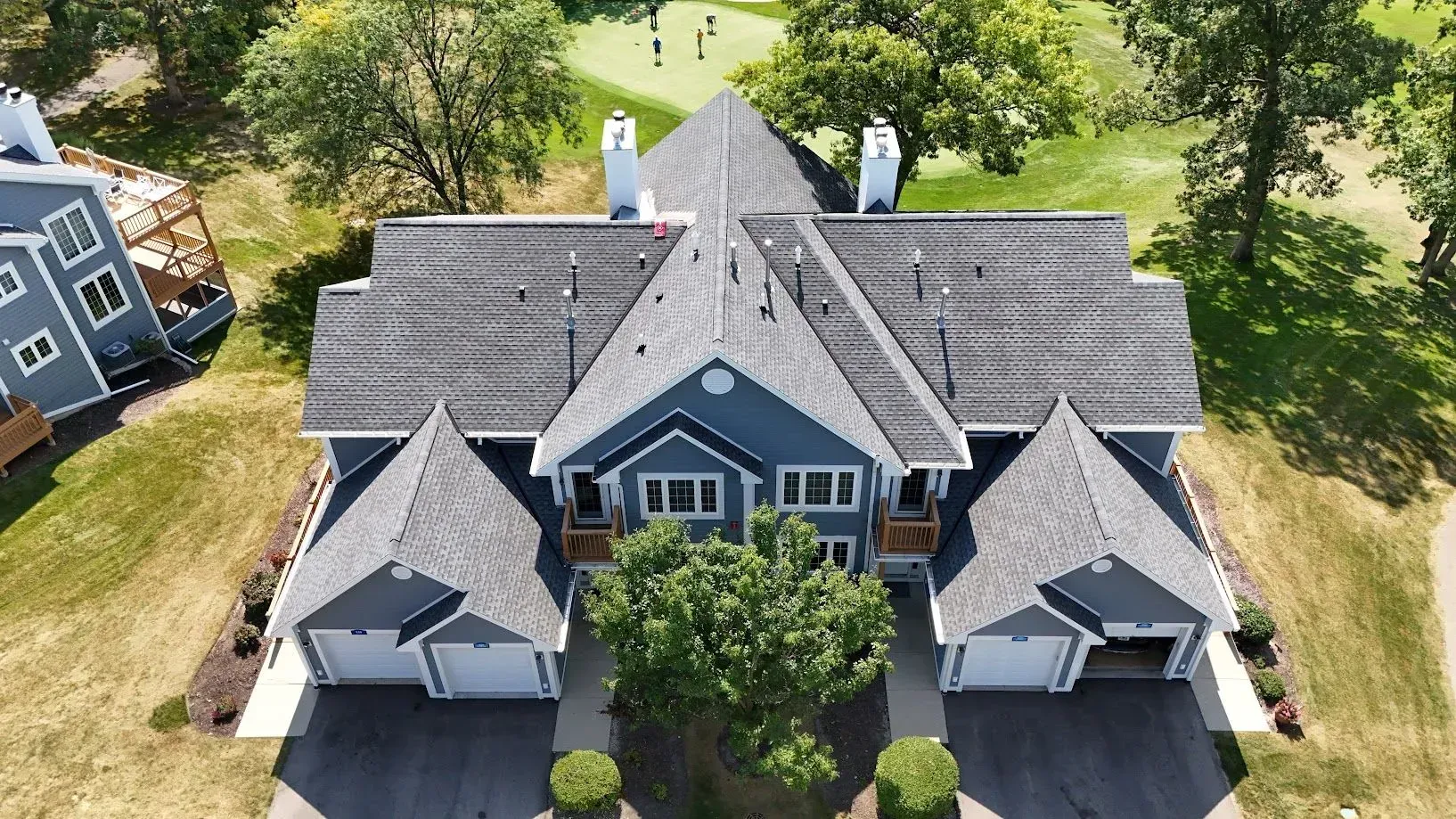 Aerial view of a blue residential building with a gray roof and two garages. A golf course is in the background. Aerial view of a large gray house with multiple gables, surrounded by trees and lawn.