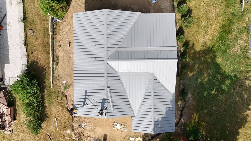 Aerial view of a house roof under construction with gray metal panels and surrounding yard.