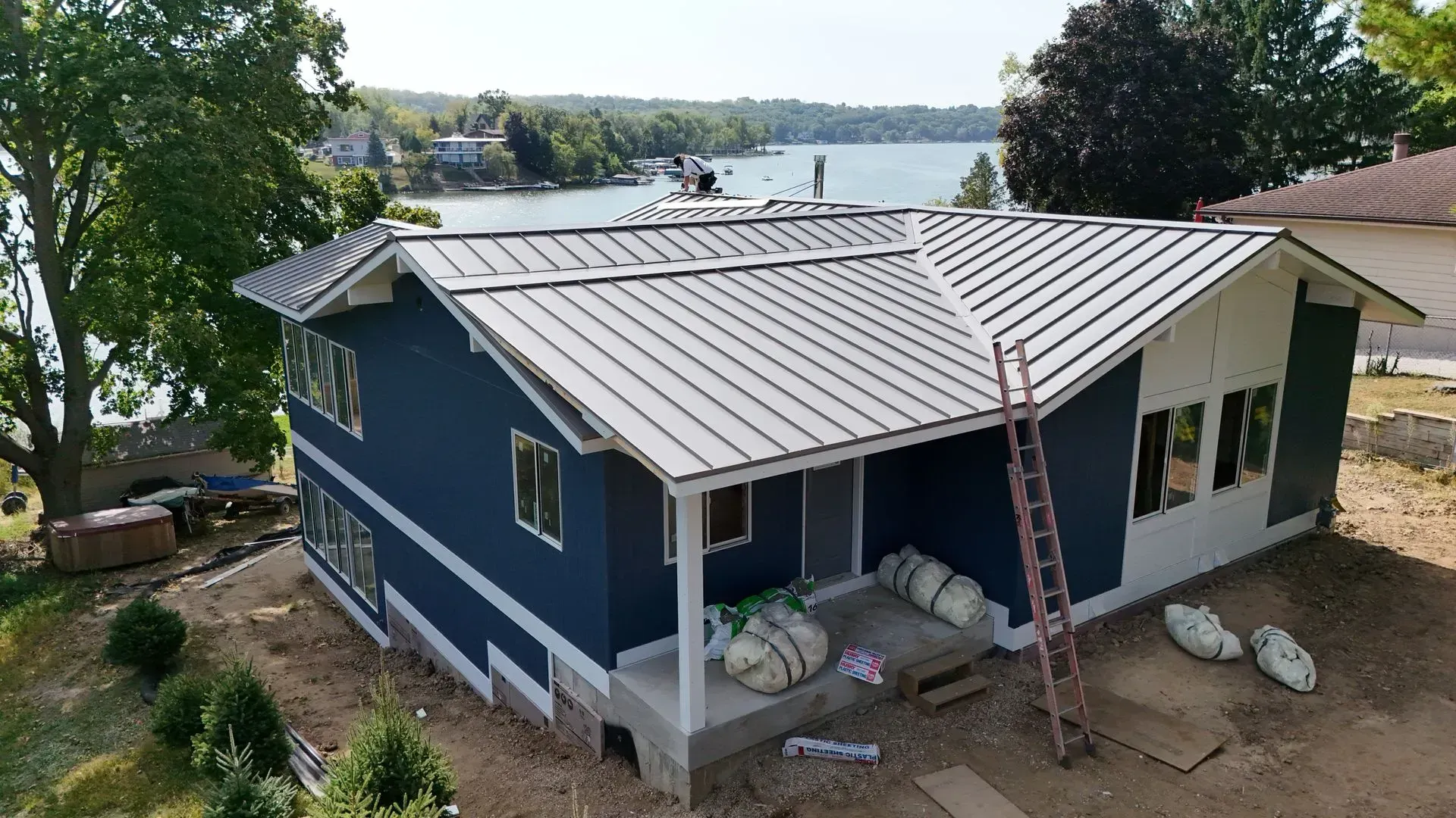 Blue house with metal roof under construction by a lake. Blue house with metal roof on a wooded lakeside lot, viewed from above.
