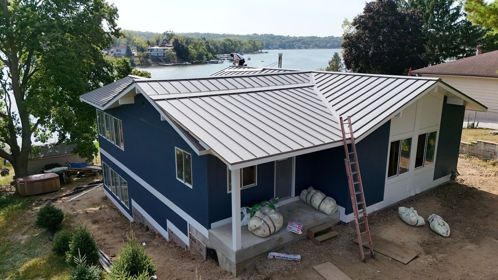 Blue house with metal roof beside a lake, with a ladder leaning against the side during exterior work