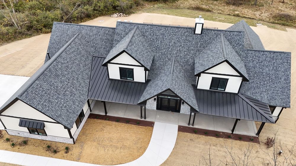 Aerial view of a large gray-roofed house with white trim and a curved driveway in a dry yard
