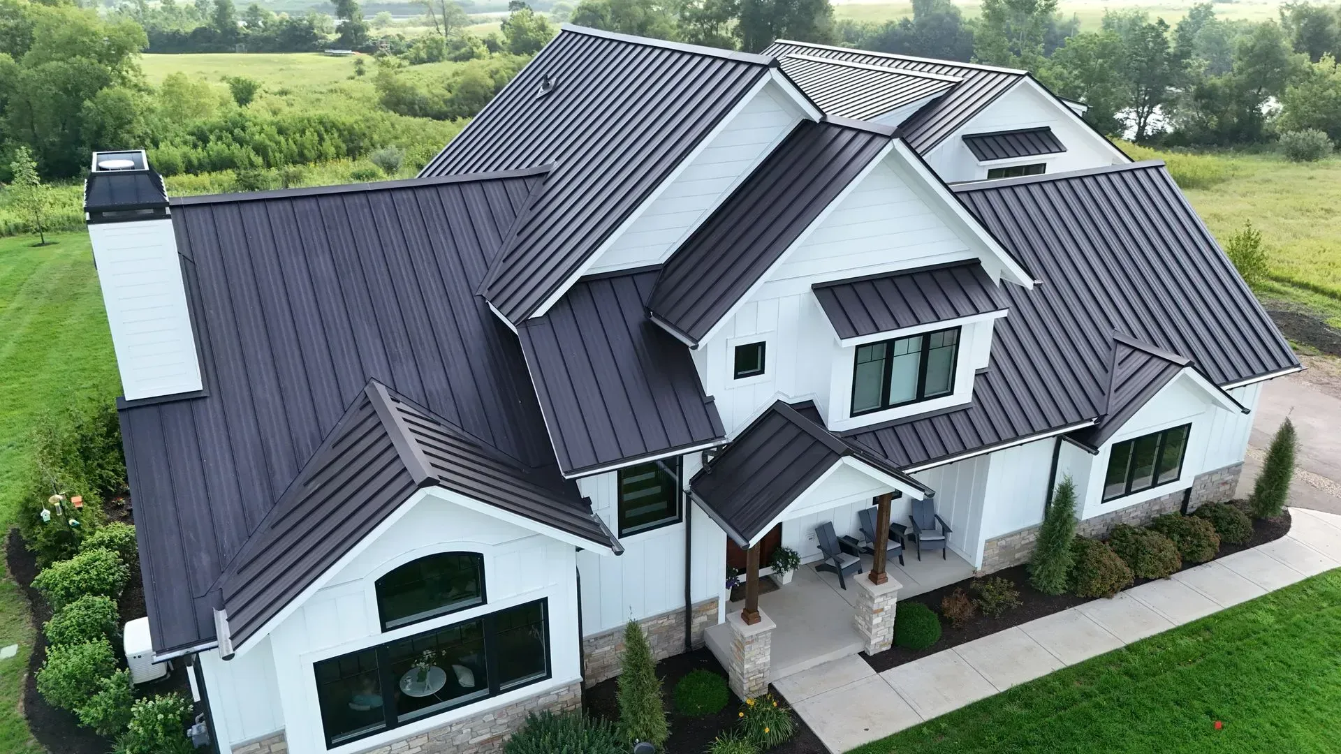 White farmhouse with dark gray metal roof, chimney, and stone accents, set in a green field. Aerial view of a modern white house with dark gray roof and front porch, surrounded by green lawn.