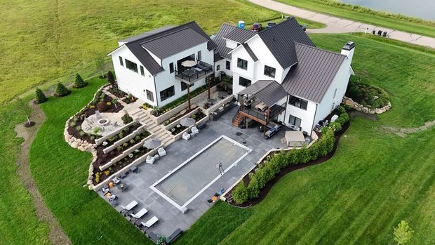 Aerial view of a modern white house with a landscaped yard, patio, and tennis court surrounded by green fields