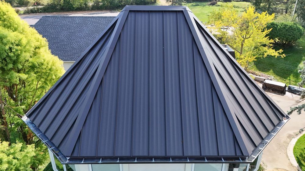 Black metal roof with steep gabled peak on a house, surrounded by trees and greenery.