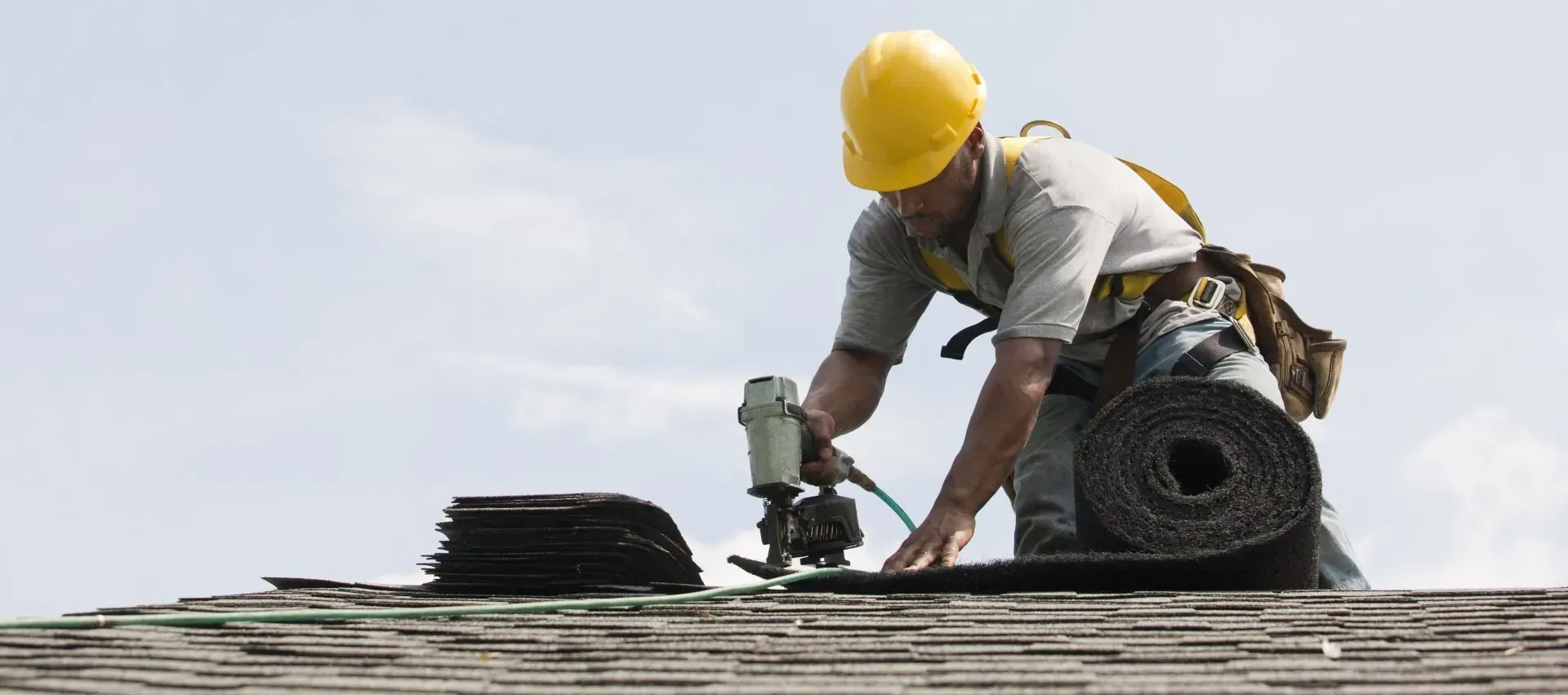 Roofer in yellow hard hat uses a nail gun to install shingles on a rooftop. Worker in a yellow hard hat kneels on a rooftop, using a power tool near rolled roofing materials.