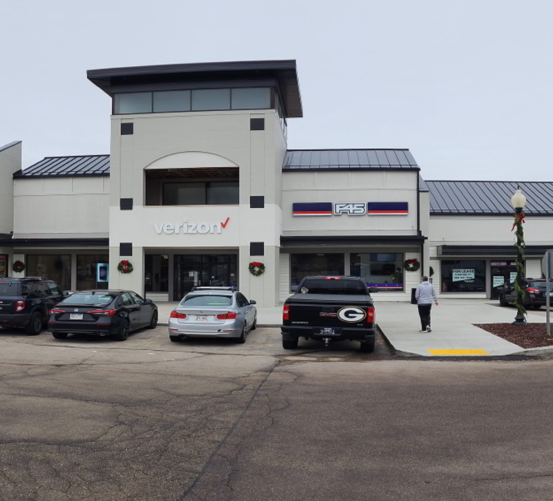 Strip mall storefronts with parked cars and a person walking on the sidewalk