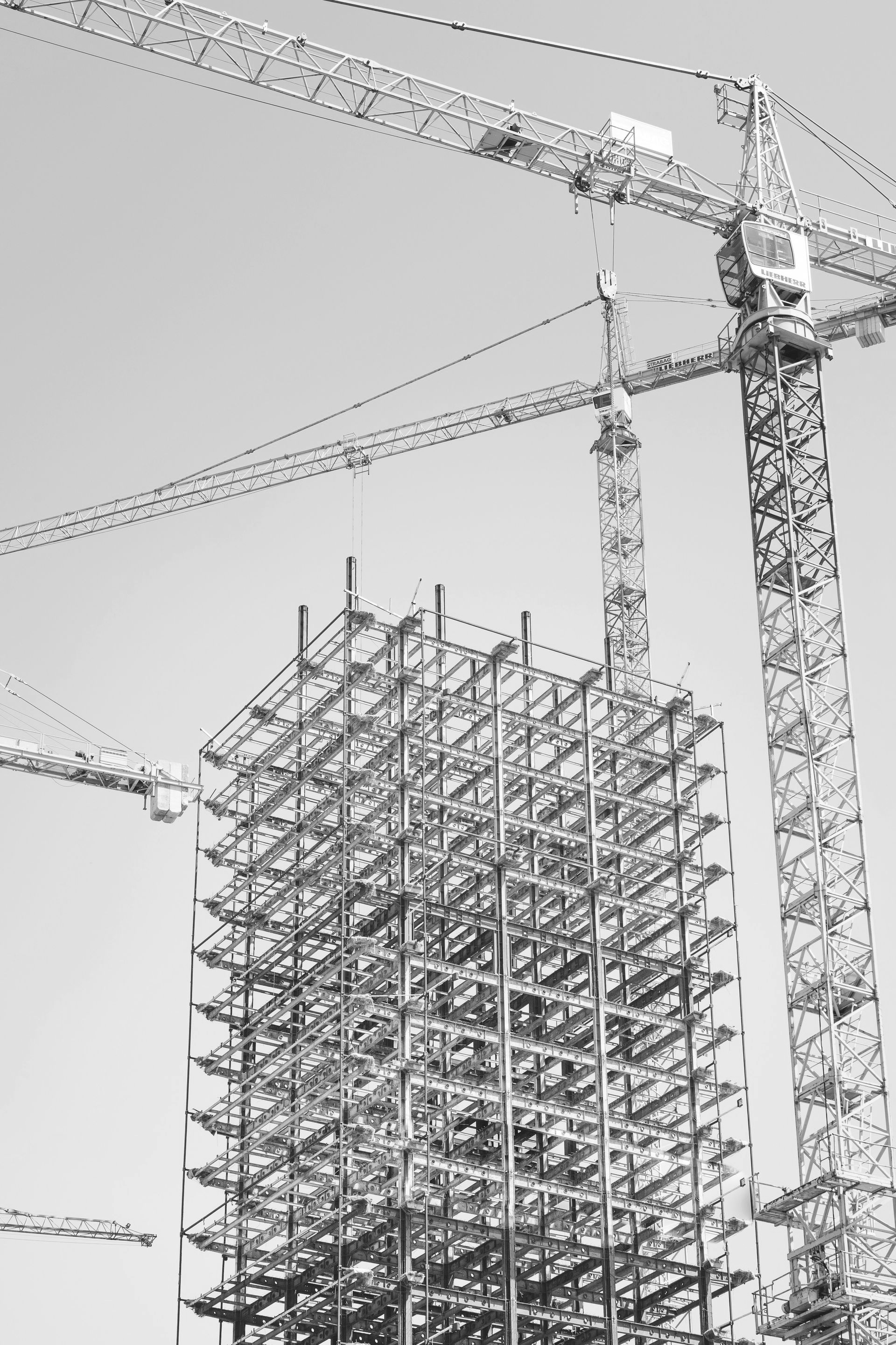 Steel frame of a tall building under construction, with several yellow cranes against a blue sky.