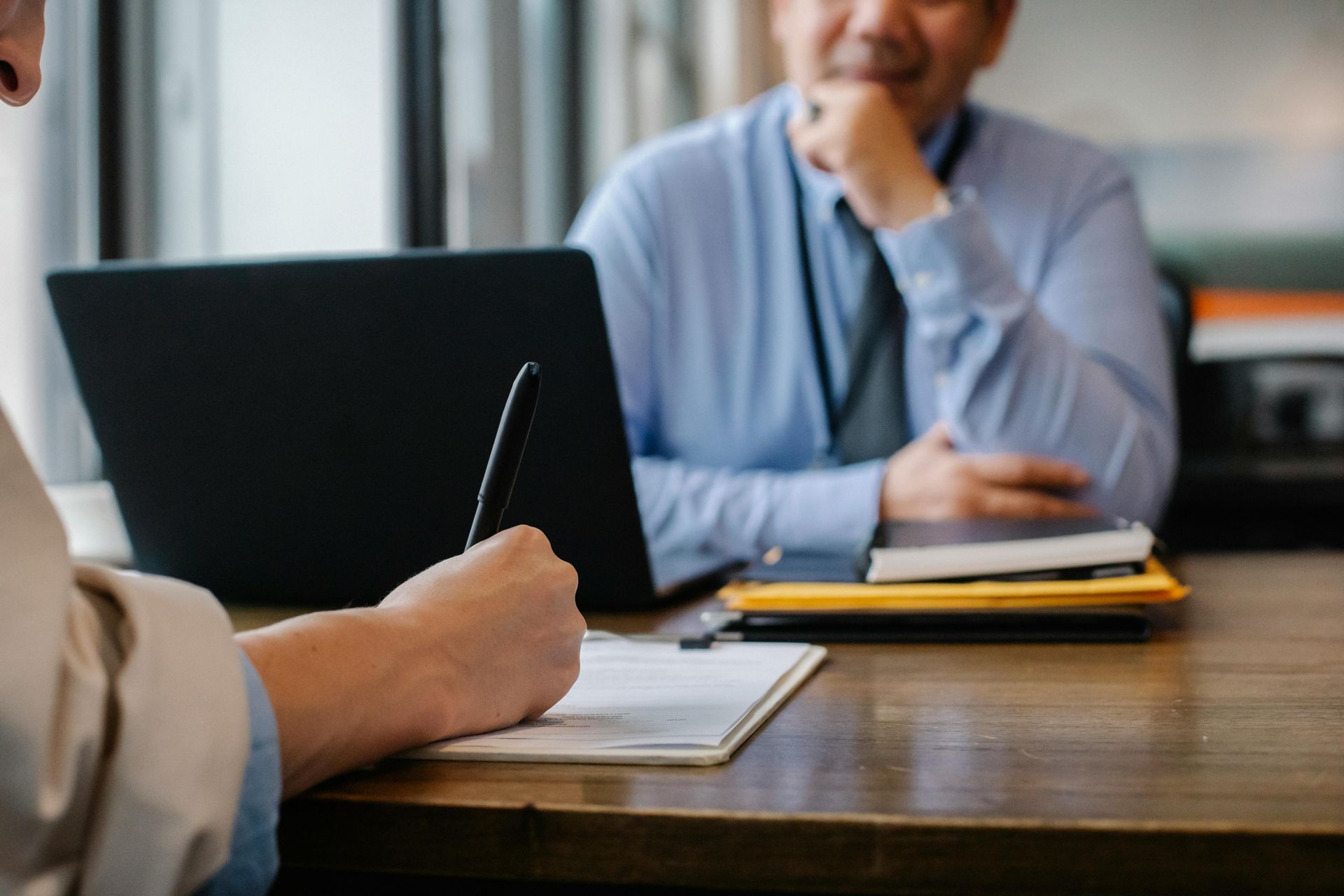 Person writing on a clipboard across from a person in a blue shirt, at a table with a laptop.