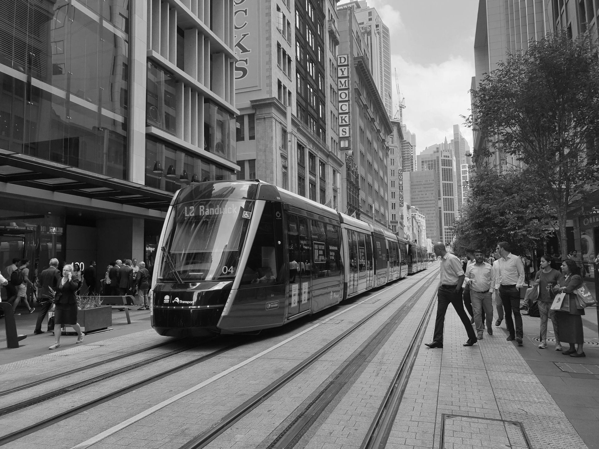 A red and black light rail train on tracks in a city street. People walk nearby.