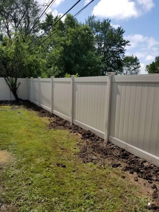 White vinyl fence along a grassy yard, dirt path at base, trees, blue sky.