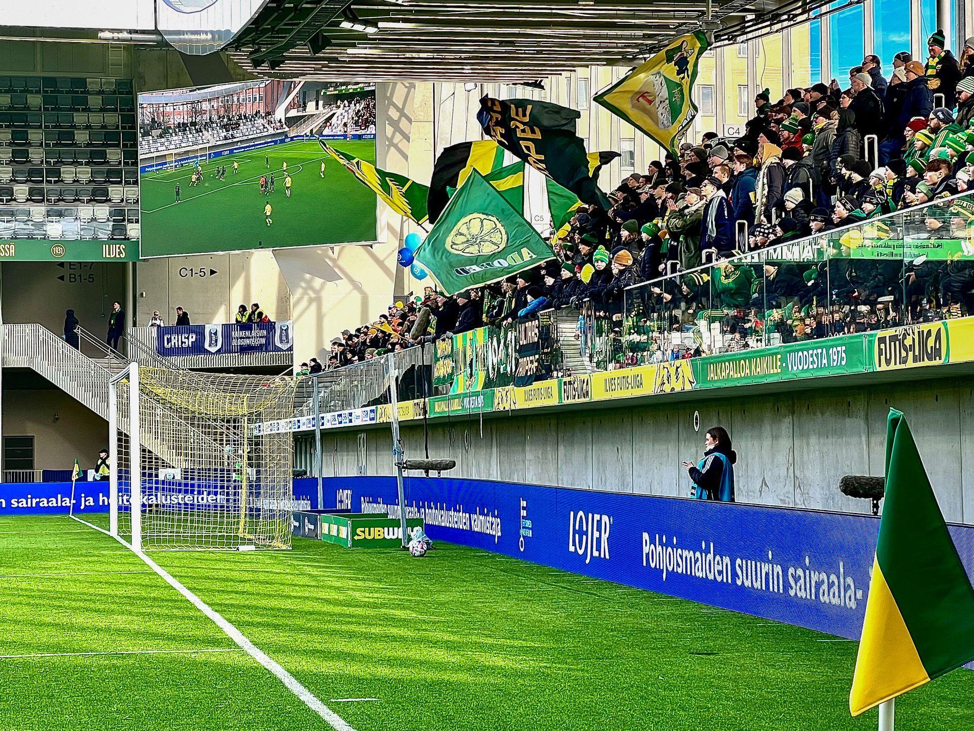 Jalkapallostadion, jossa vihreisiin ja keltaisiin pukeutuneet fanit hurraavat. Kenttä, pelaajat, liput.