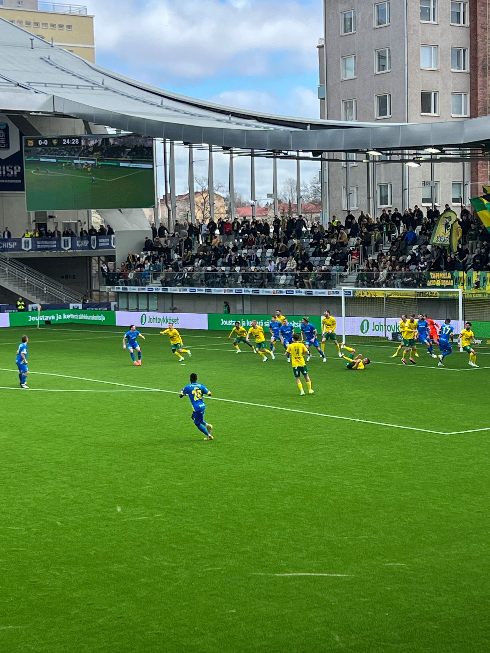 Jalkapallo-ottelu stadionilla. Keltaiset ja siniset joukkueet vihreällä kentällä. Katsojat katsomossa katsovat ottelua.