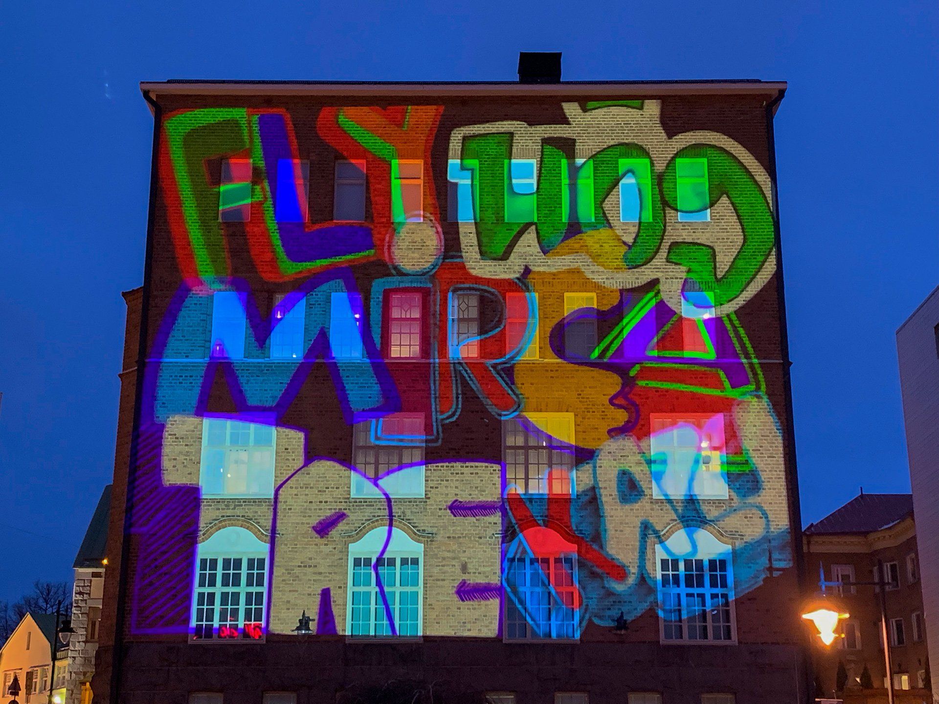 Värikästä graffititaidetta heijastettuna tiilirakennukseen hämärässä. Mukana teksti 
