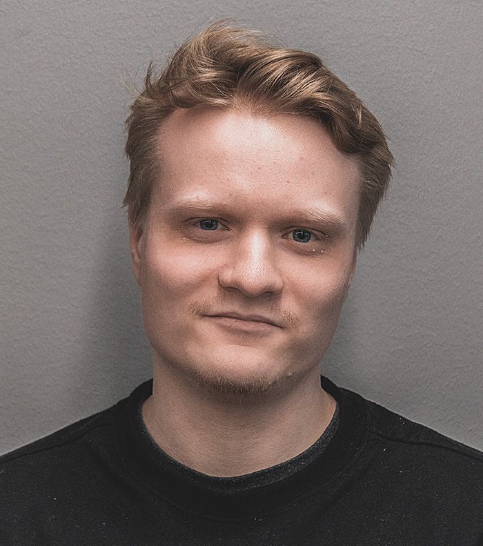 Vaaleatukkainen mies, jolla on neutraali ilme ja musta paita, harmaata taustaa vasten.