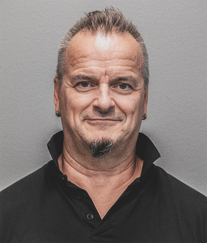 Harmaantuvat hiukset ja pukinparta, musta paita yllään, hymyilevä mies.