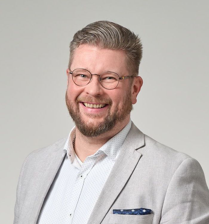 Silmälasit ja bleiseri päässä oleva hymyilevä mies. Vaalea tausta.