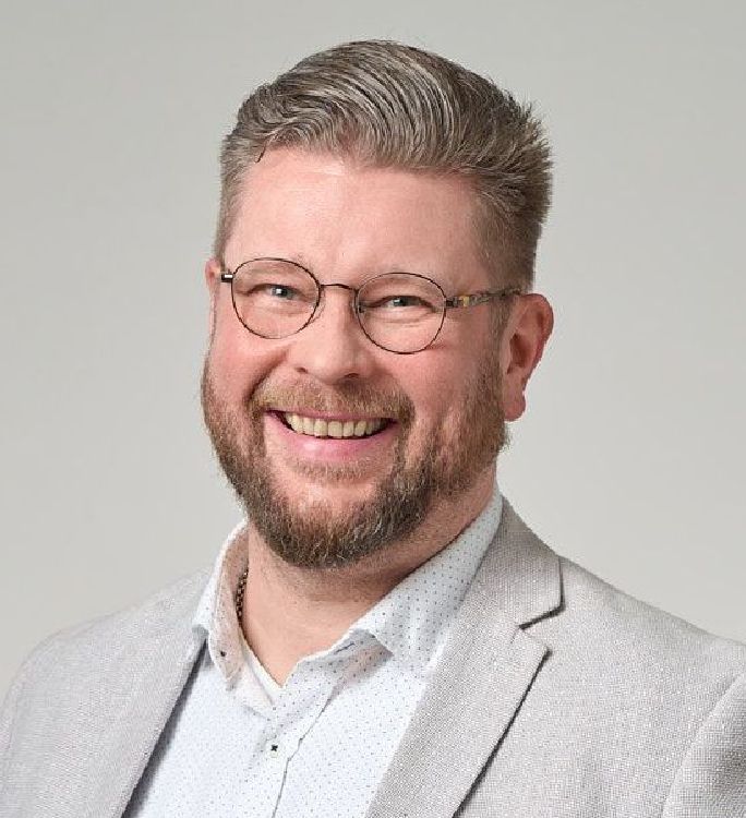 Silmälasit ja parrakas hymyilevä mies, jolla on yllään vaaleanharmaa bleiseri ja kauluspaita.