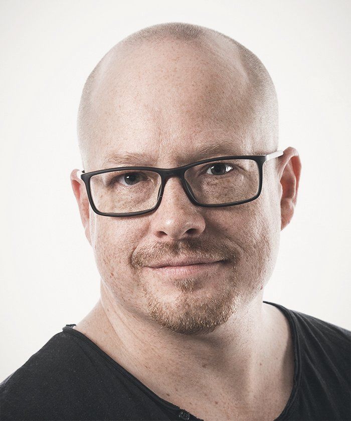 Silmälasipäinen, kaljuksi ajeltu mies hymyilee ja katsoo eteenpäin. Hänellä on yllään musta paita valkoista taustaa vasten.
