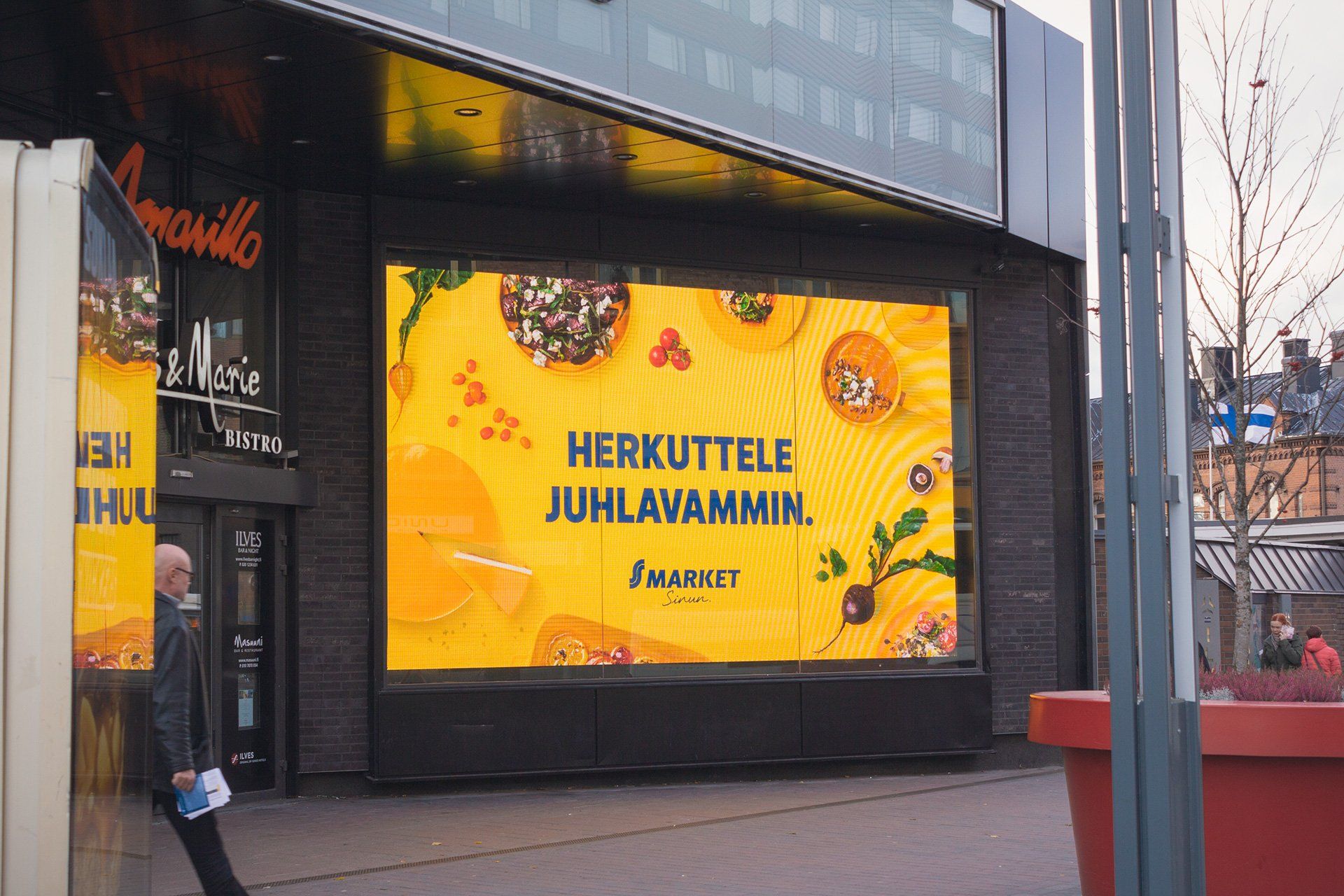 Suuri digitaalinen ruokaa mainostava kyltti, teksti suomeksi, ravintolan sisäänkäynnin vieressä.