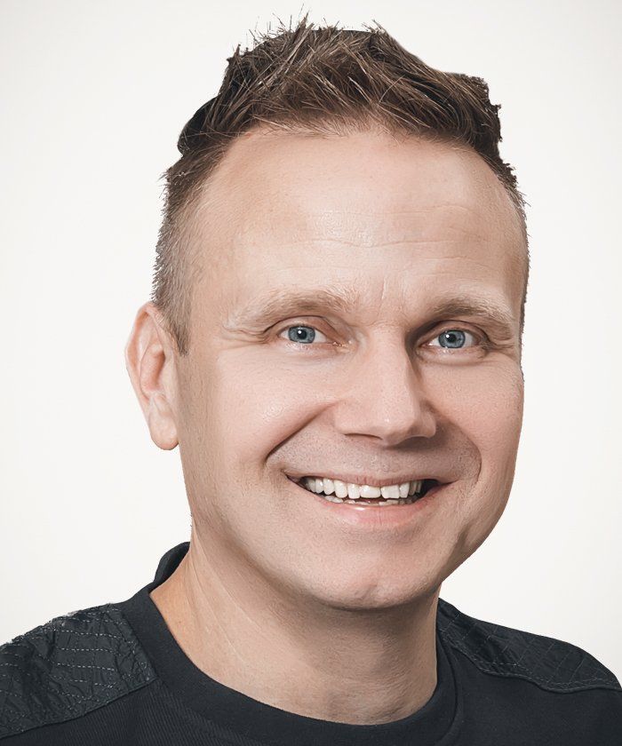 Lyhyt, piikkinen ruskea tukkainen mies hymyilee leveästi ja katsoo suoraan katsojaa.