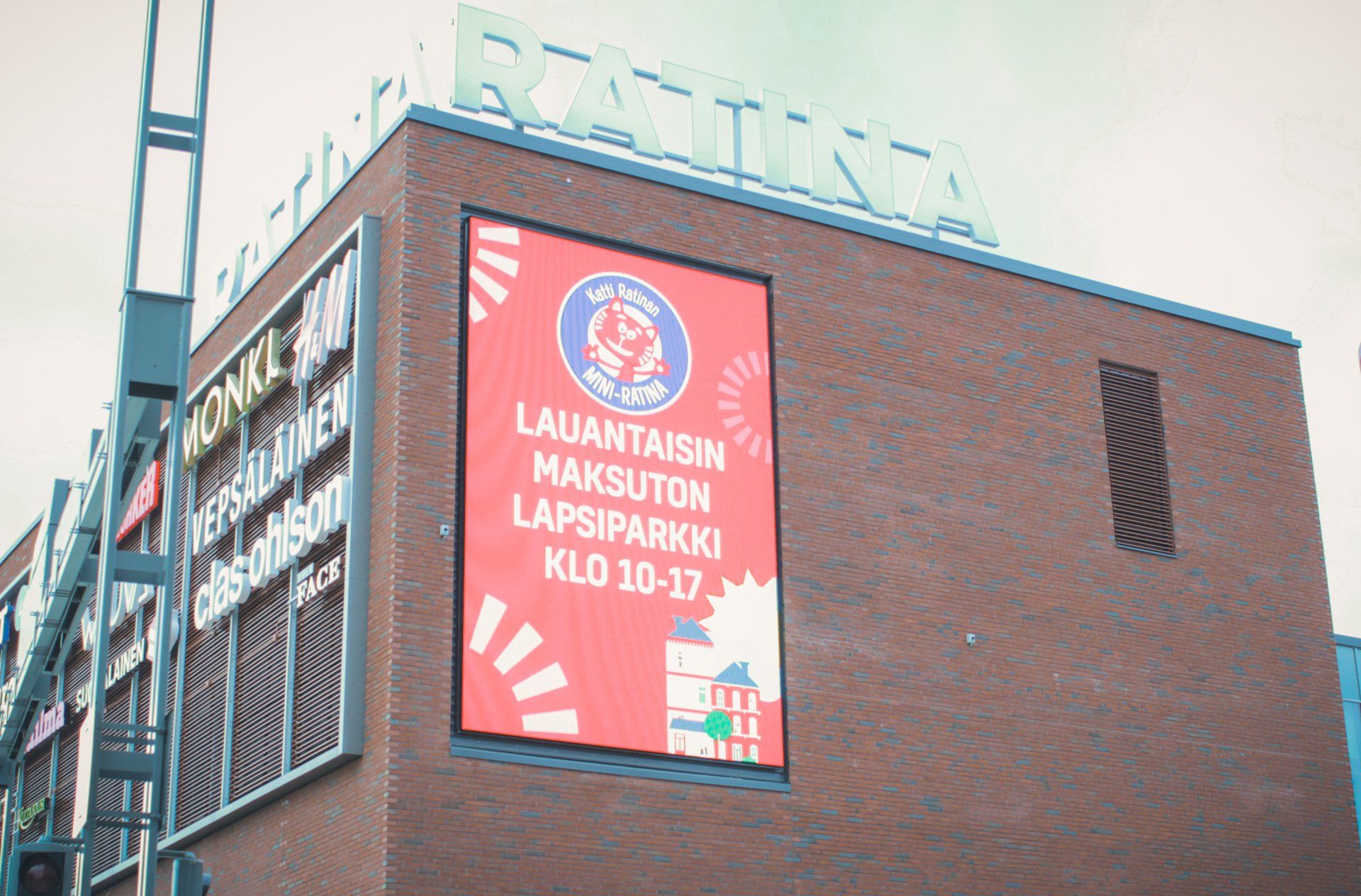 Rakennus kyltillä: Ilmainen lasten leikkialue lauantaisin klo 10–17. Punainen, sininen logo ja teksti.
