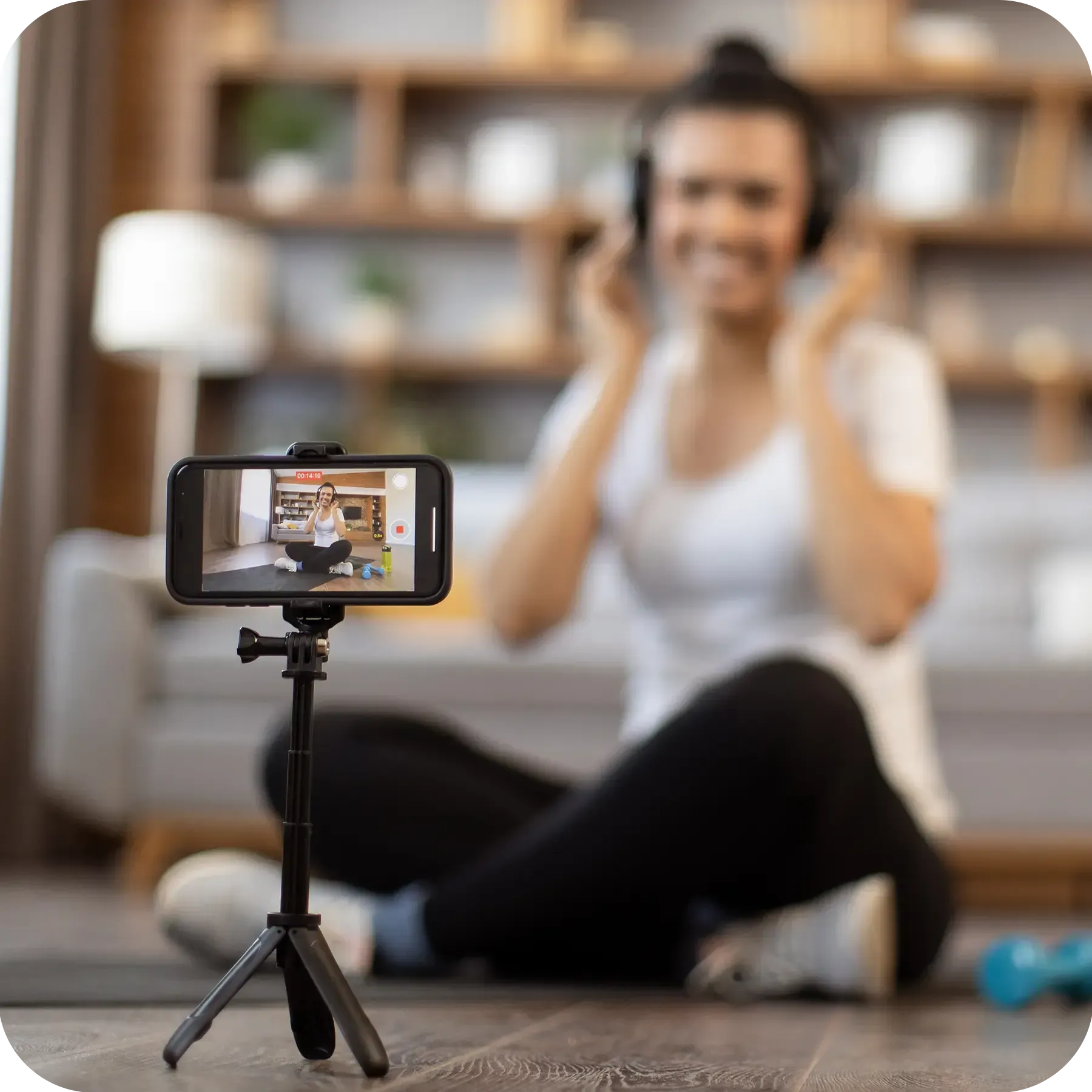 Healthy fit multiracial woman sitting in cross-legged pose with headphones on while recording video content of fitness training for online sharing