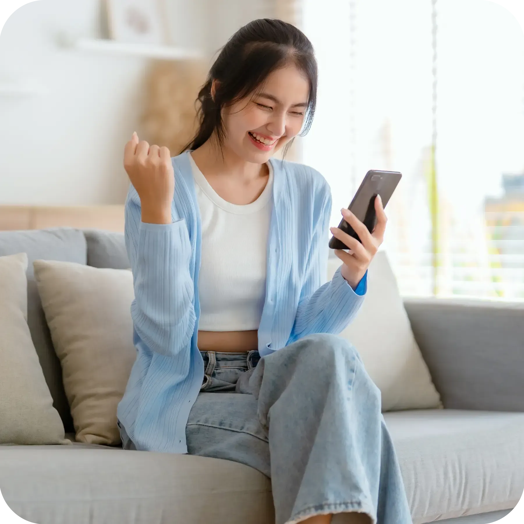 Smiling pretty young Asian woman sitting on cozy couch holding her phone