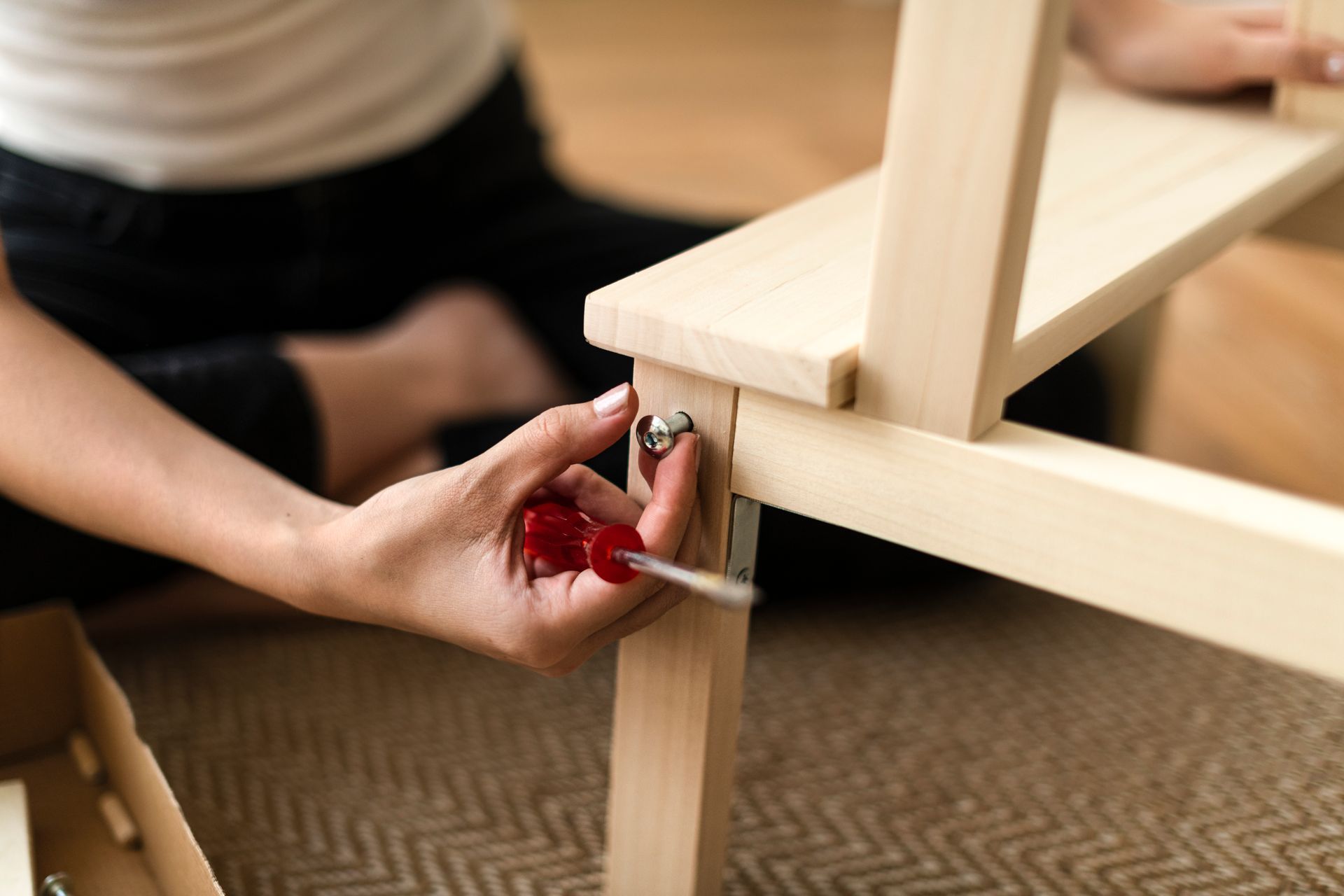 A person uses a red-handled screwdriver to assemble a light-colored wooden piece of furniture.