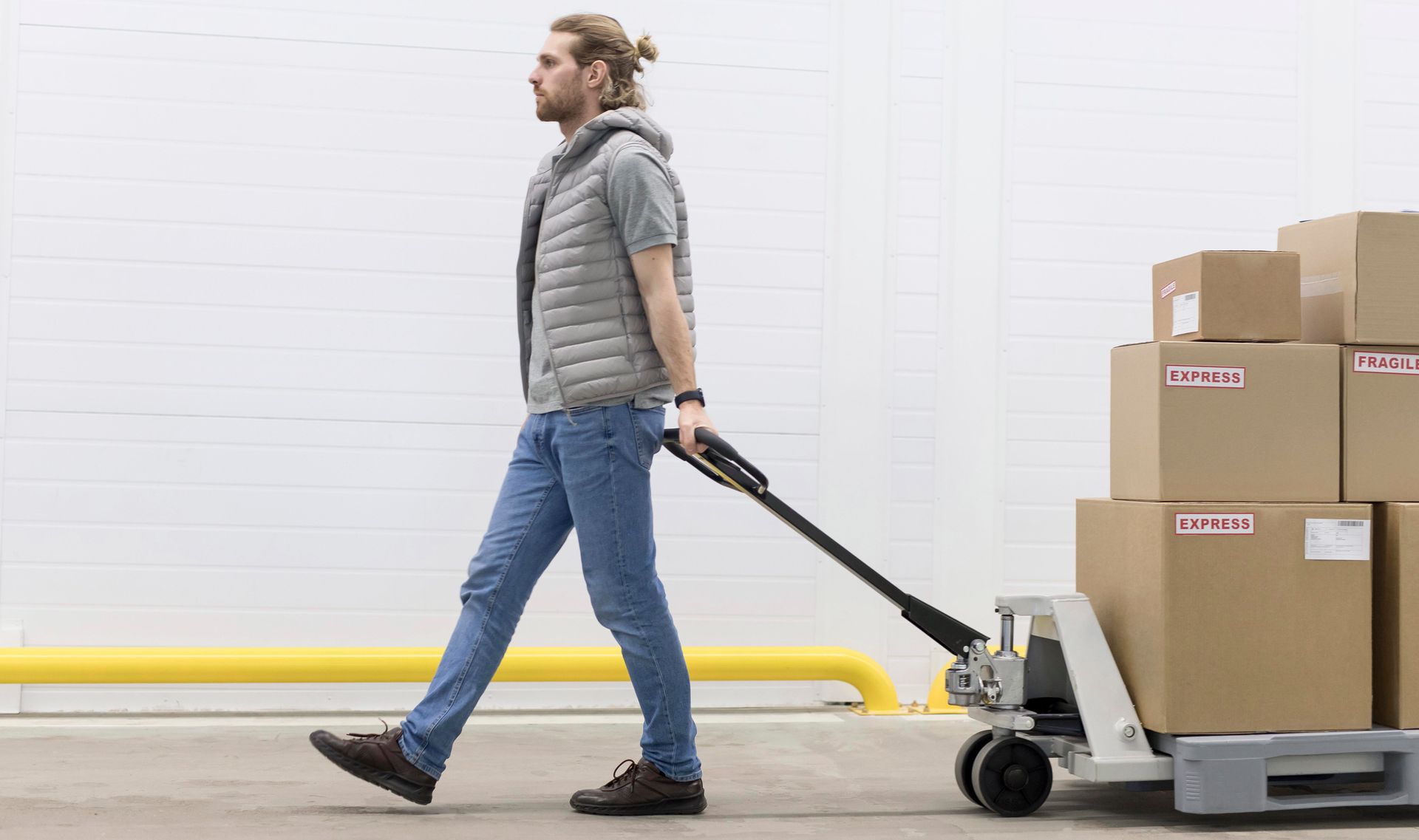 Man pulling a pallet jack loaded with cardboard boxes through a warehouse aisle