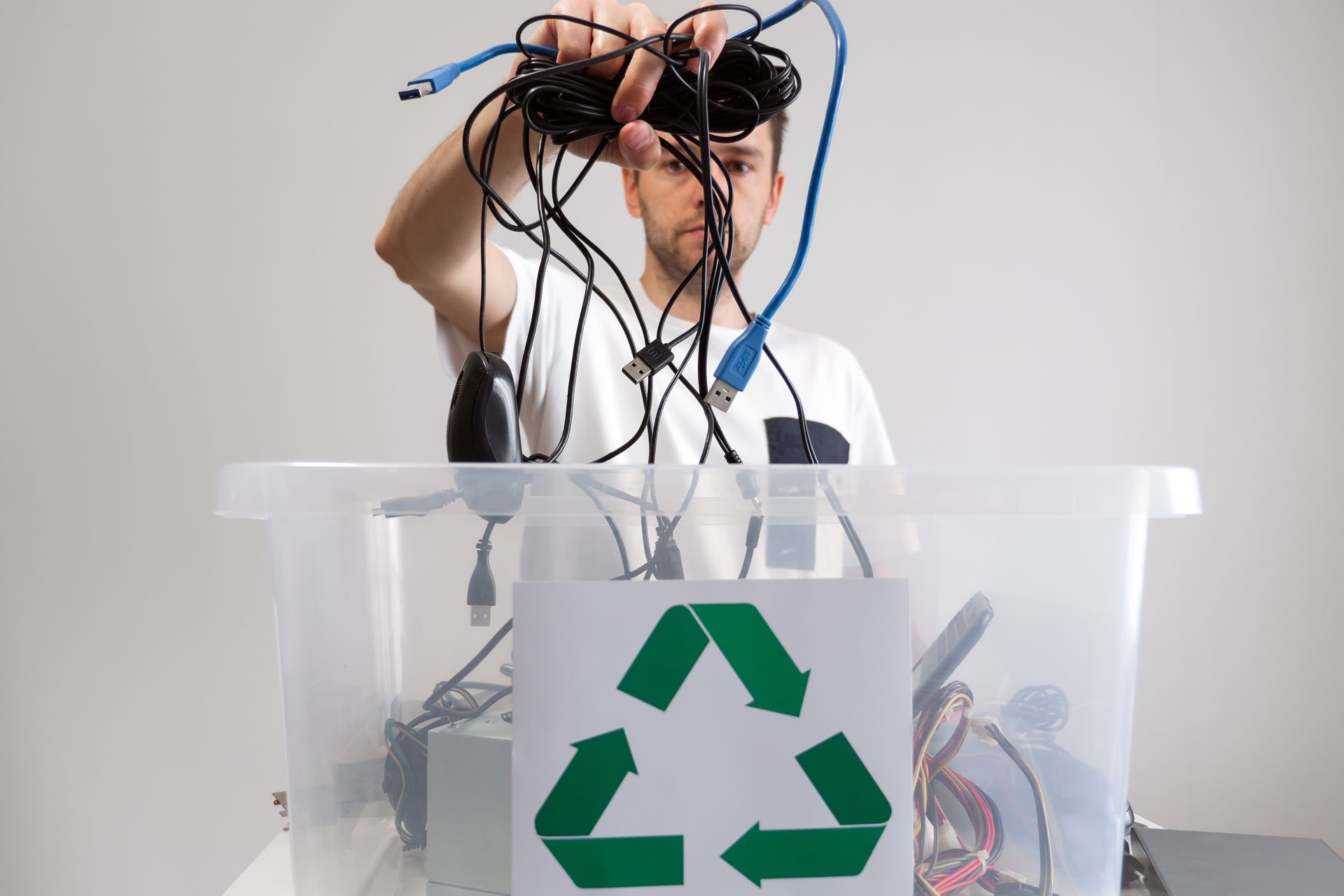 Person holds tangled cables over a clear recycling bin with a green recycling symbol.