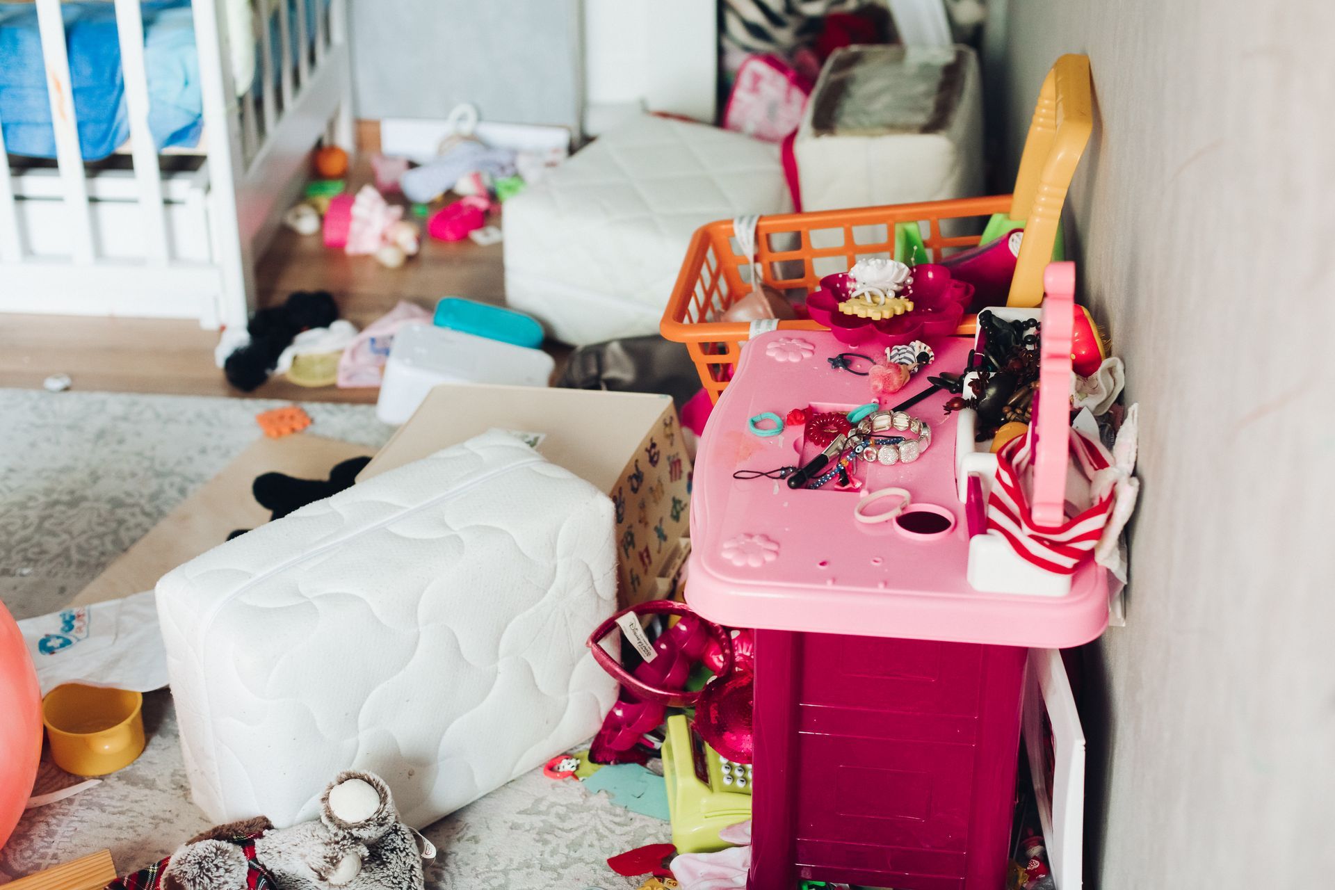 Disordered playroom with overturned white ottoman, scattered toys, and pink storage bin cluttered with items