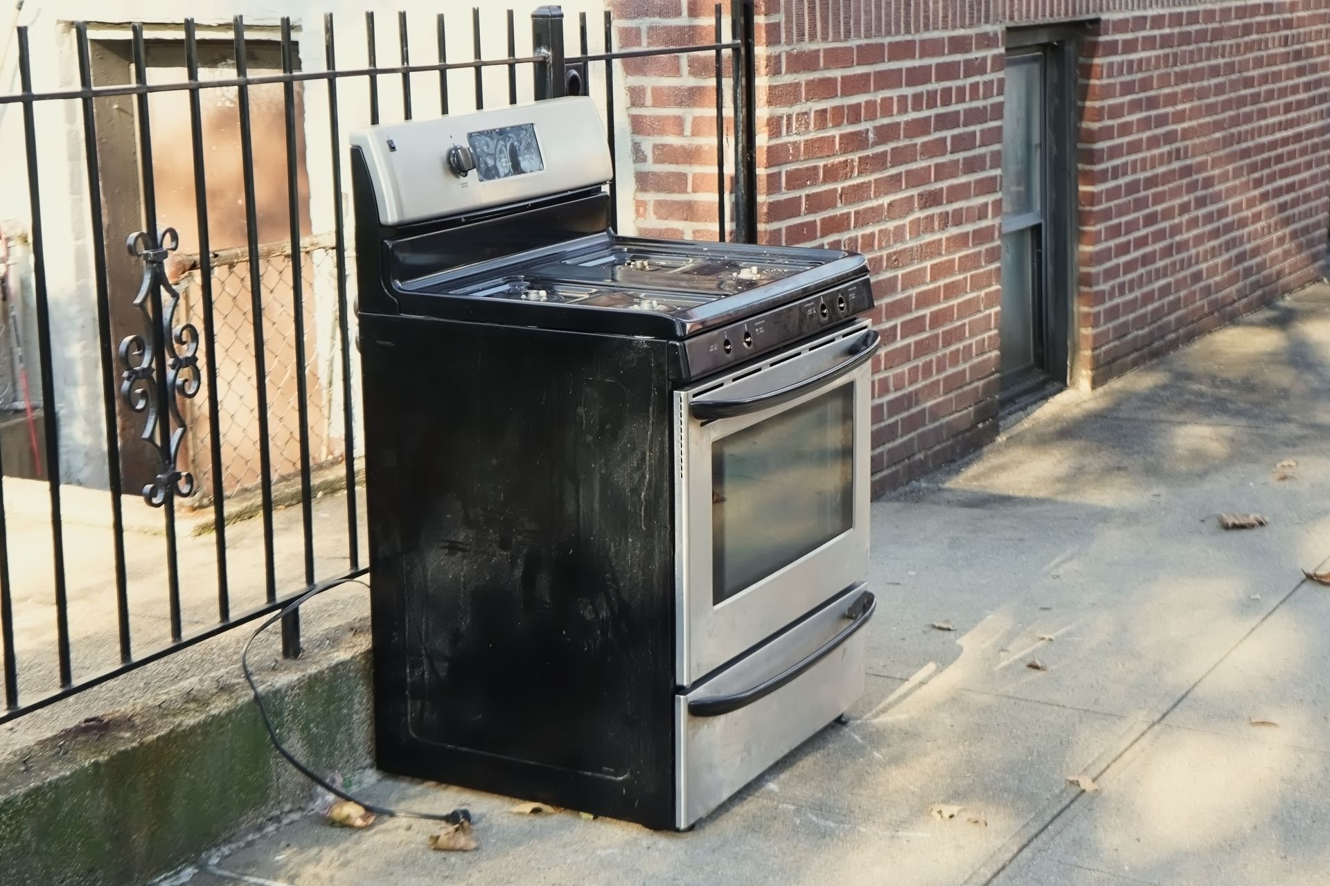 Black-and-silver stove sitting on a sidewalk beside a brick wall and iron fence.