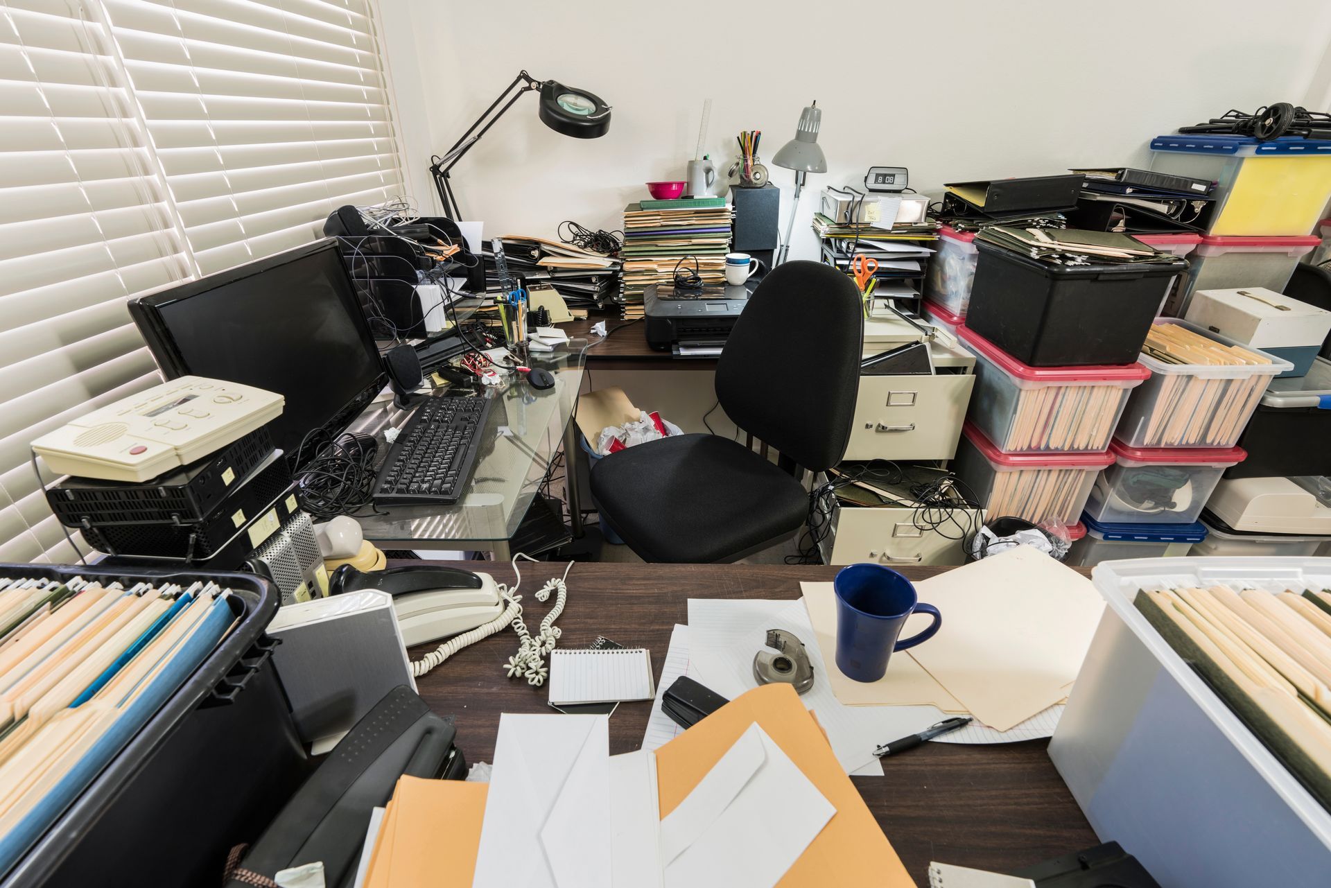 A messy office with a desk , chair , computer , and lots of papers.