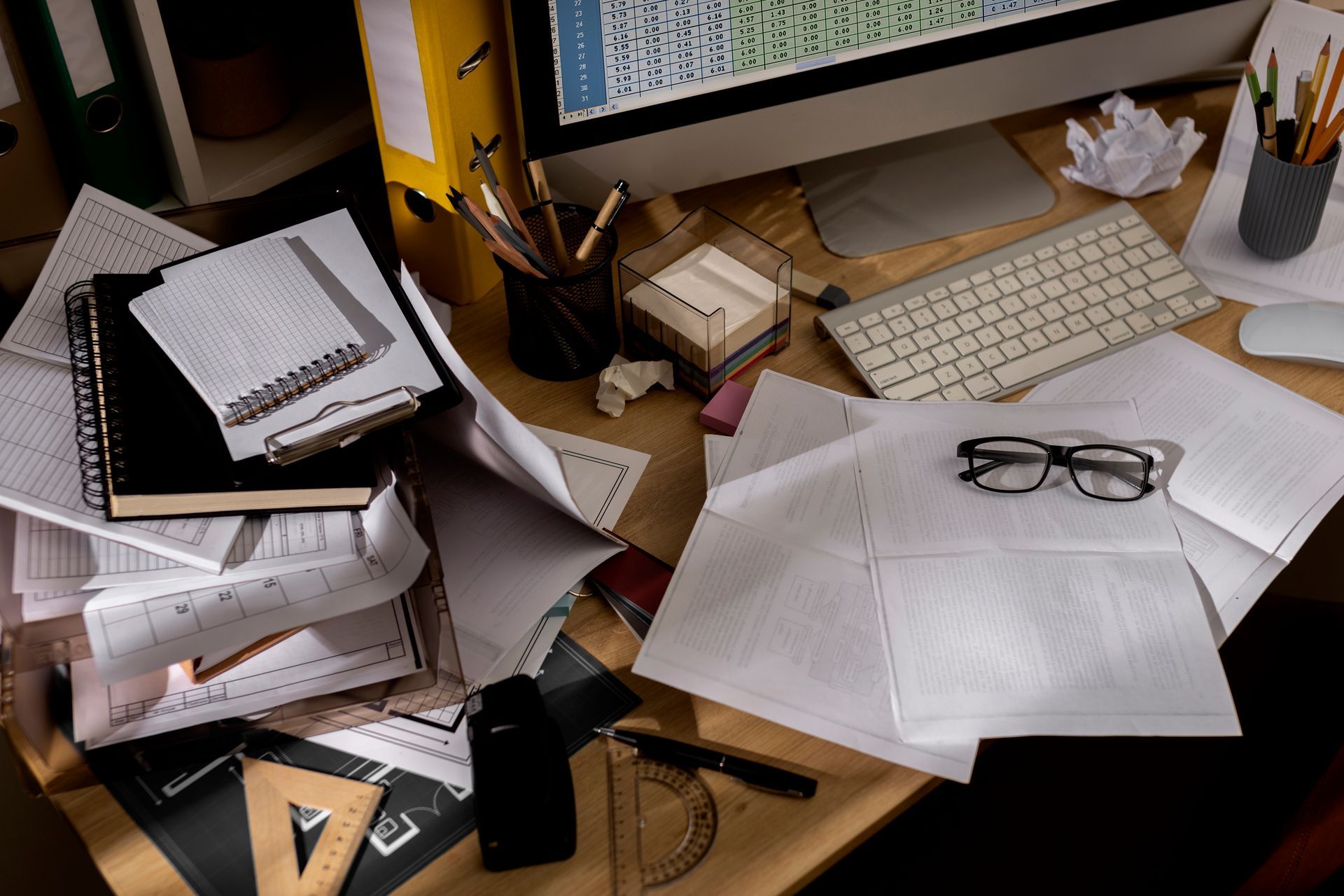 A messy desk with a computer , notebooks , glasses , and a stack of papers.