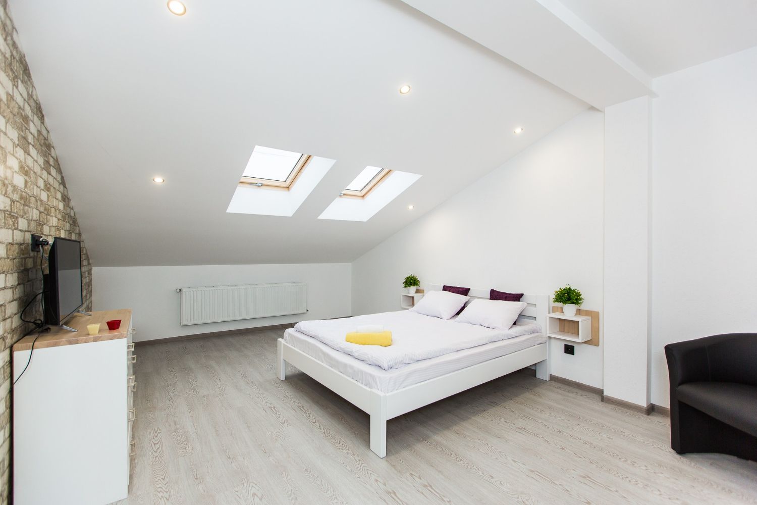 Bright white attic bedroom with skylights, low bed, TV, and black armchair