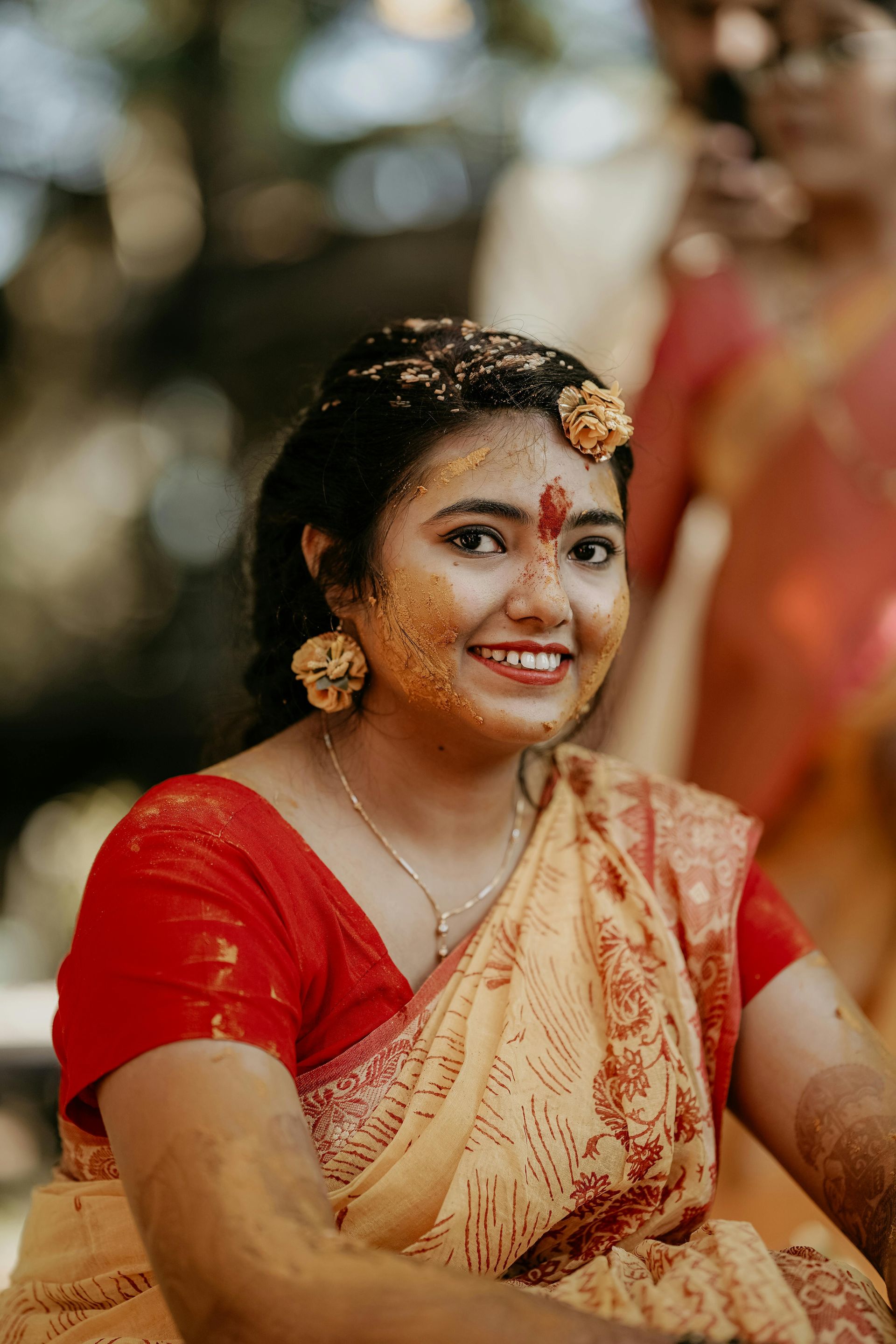 A woman in a red blouse and a yellow saree is smiling.