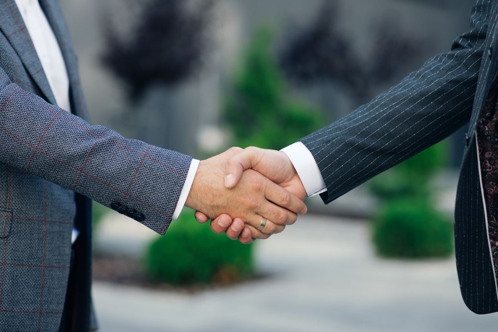 Two people in suits shaking hands outdoors in front of a blurred building and greenery
