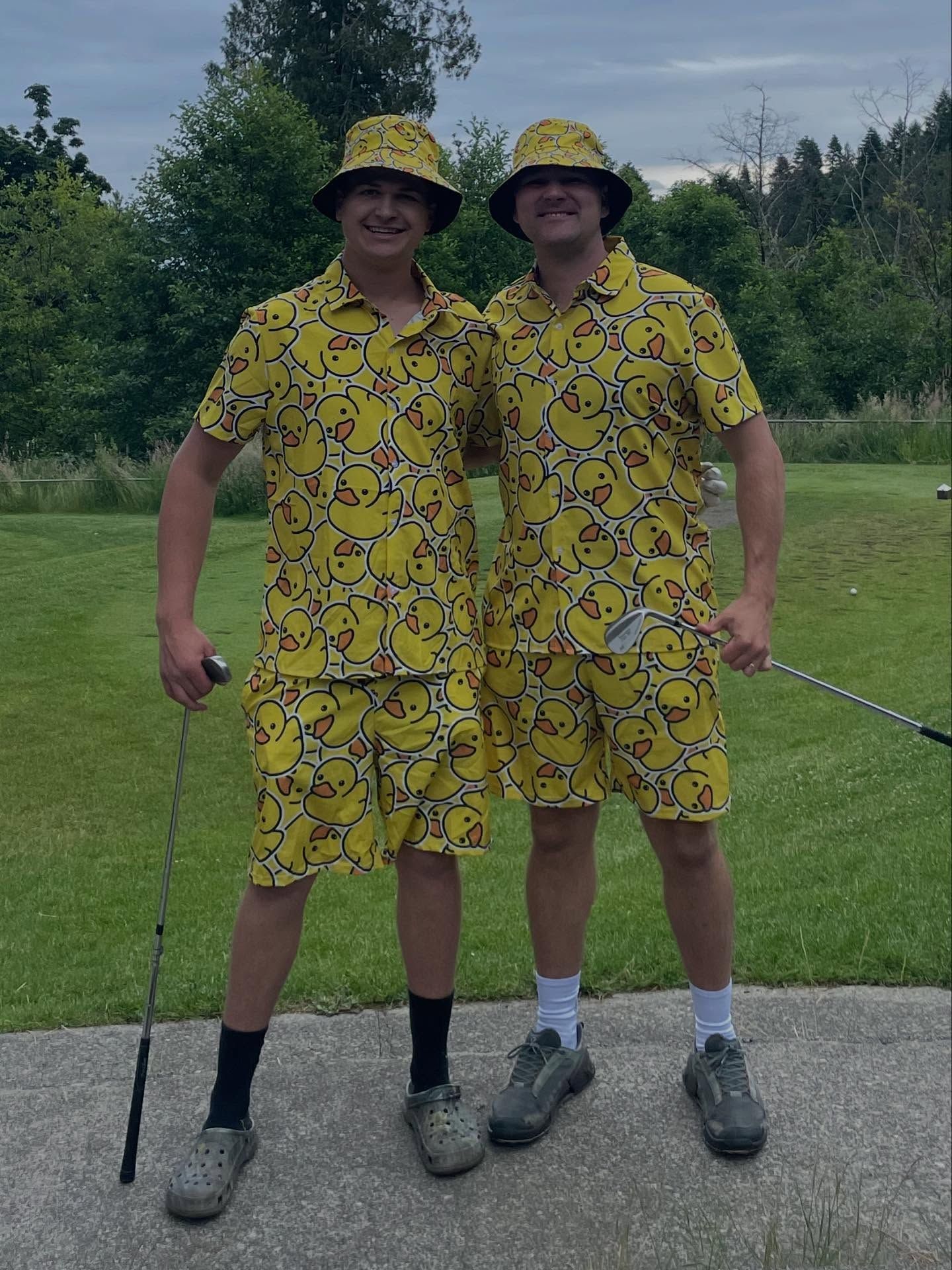 Two golfers in matching yellow outfits stand on a path, holding clubs on a grassy course.