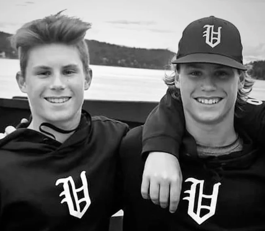 Two smiling friends in black hoodies and cap, posing by the water with arms around each other
